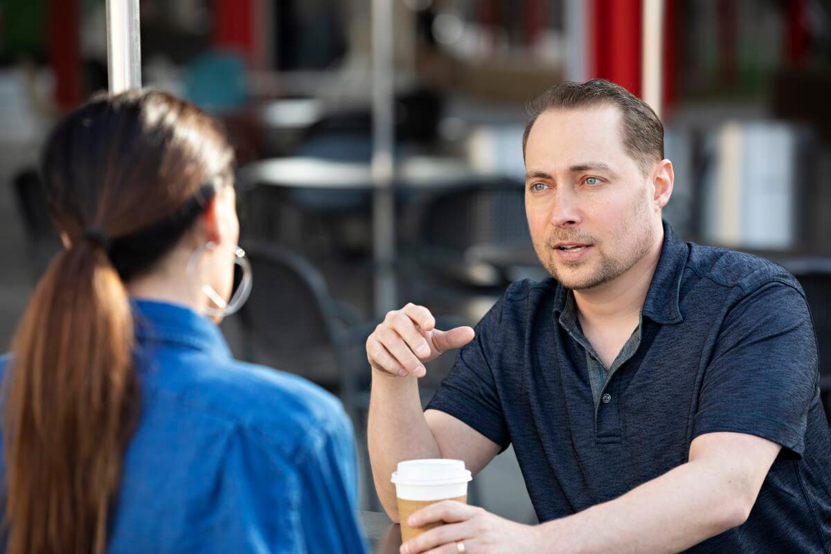 A picture of Business Coach Dave Labowitz in a coaching session. He is on the right, with a dark blue, collared, short sleeve shirt. He's holding a cup of coffee on a table in front of him with his left hand and gesturing with his right hand. There is a woman across from him wearing a royal blue shirt. She is facing away from the camera so you can only see is her long, straight brown hair.