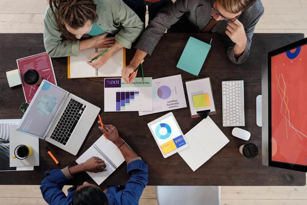 A group of three office-workers sitting a conference table, working on a digital marketing campaign. The table has an assortment of laptops, notebooks, sticky notes, and graphs/charts of digital marketing campaign metrics.