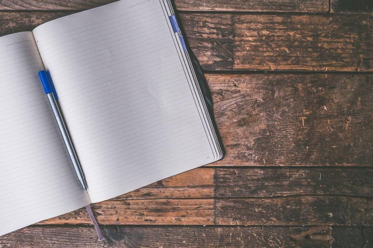 a picture of a notebook sitting on a wooden table. The notebook is open, with a pen sitting in the binding's crease between pages. This image is to suggest people document things better.