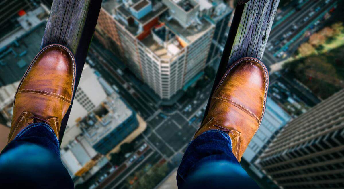 Symbolic image for fear of failure. Shows two narrow boards stretching high above a city. Viewpoint is from person balanced on those boards, looking down at their shoes, standing on the boards, with the cityscape far below.