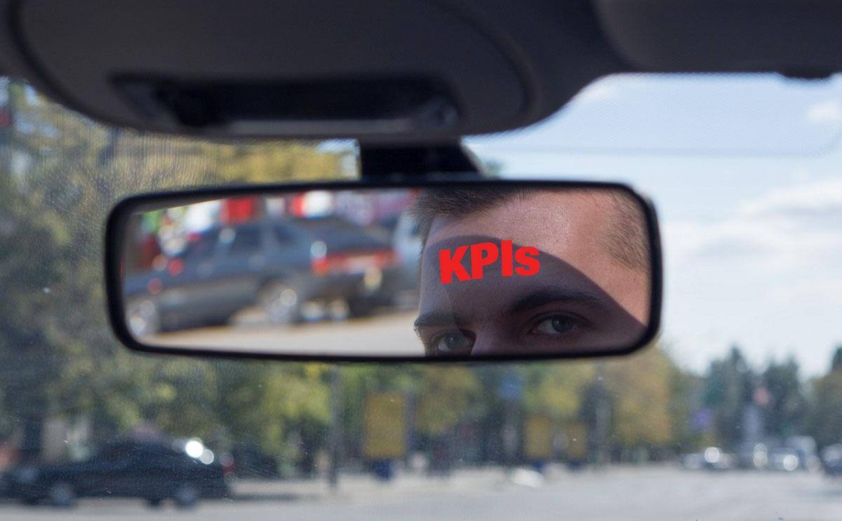 A closeup of the reflection of a man's face in the review mirror of his car. Across his forehead is the work "KPIs" in red letters.