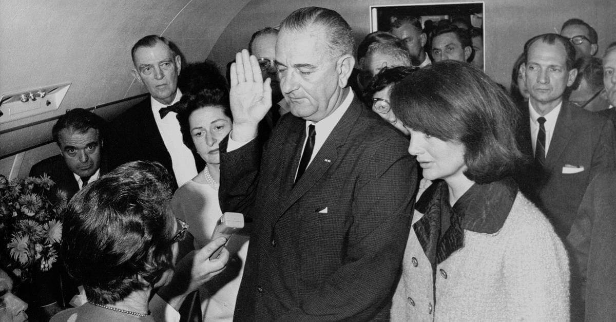 A picture of Lyndon B. Johnson swearing the oath of office for the US presidency. The picture was taken on Air Force One, shortly after JFK's death was confirmed. He is flanked on one side by his wife, Lady Bird Johnson, and on the other by Jackie Onassis, still wearing her blood-stained jacket from the day's events.