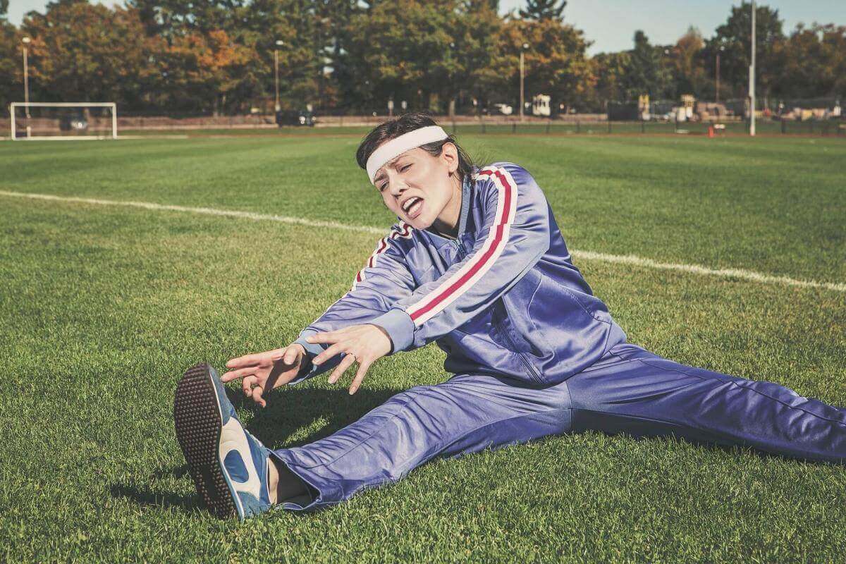 A picture of a woman wearing a blue track suit sitting on a sports field stretching her hamstrings. She looks frustrated and in pain.