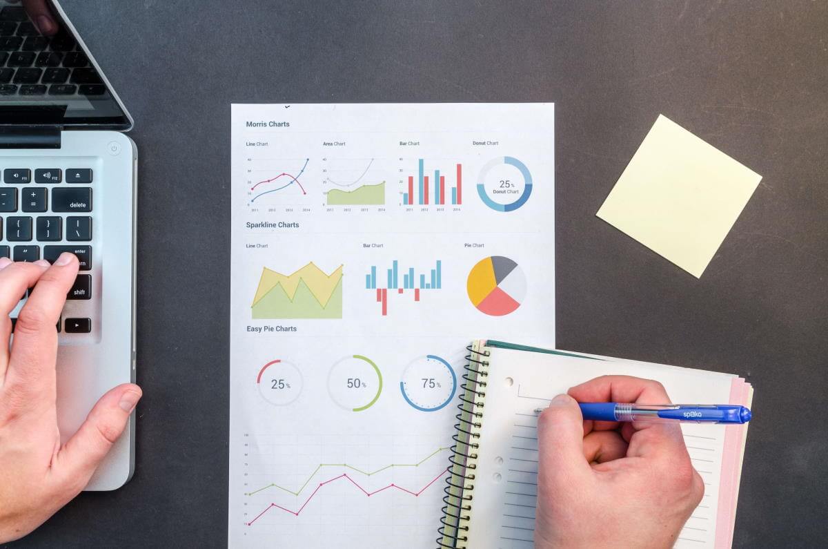 A picture of a venture-backed company's dashboard report sitting on a desk. To the left is a laptop, and overlapping the lower righthand corner of the report is a notebook. Whomever is looking at the report is taking notes in the notebook with one hand with the other hovering over the laptop.